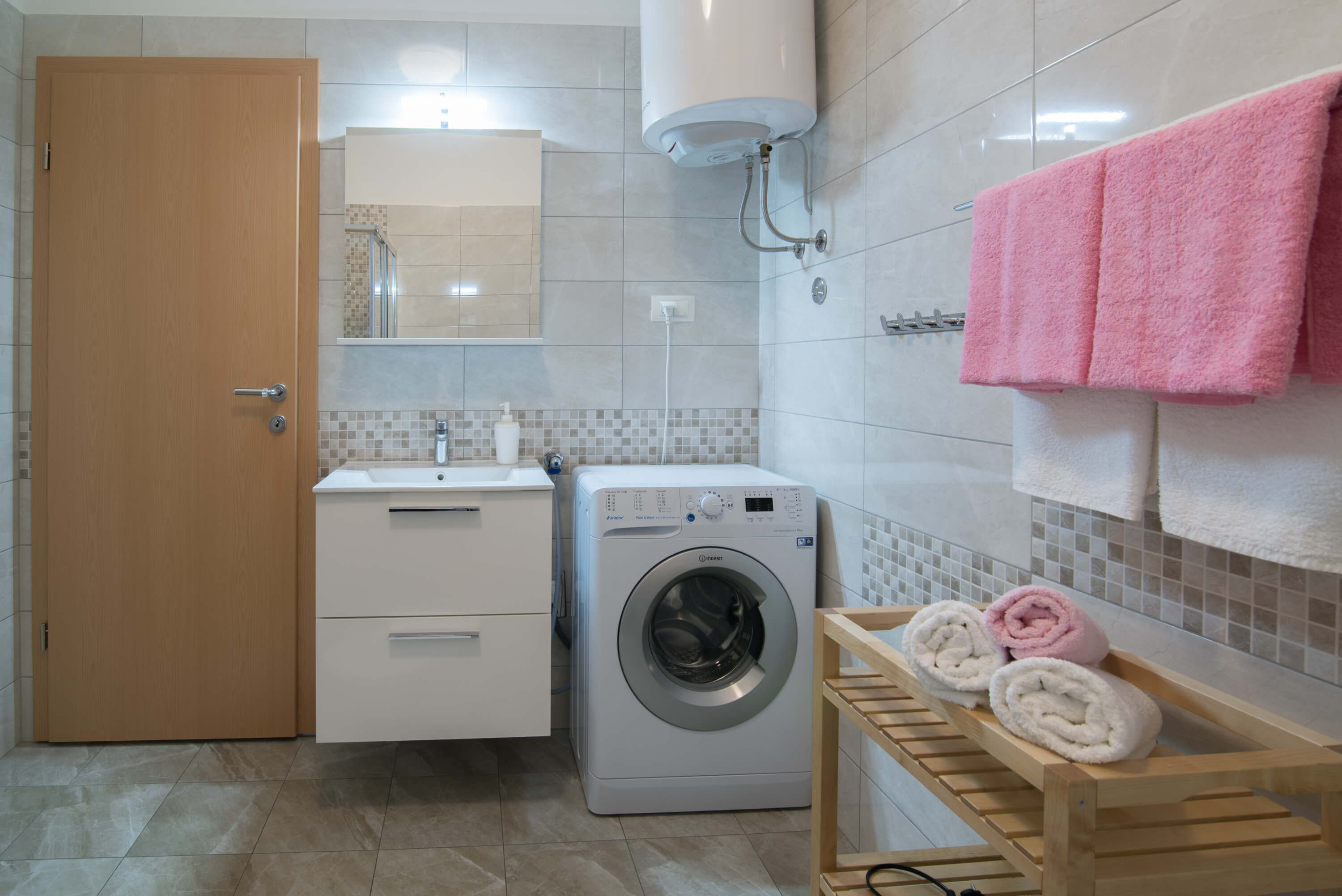 Well equipped spacious bathroom with beautiful tiles and appliances. Mirror and sink near the door. Washing machine under the boiler. Towels in different colors on the hangers and on the towel shelf.