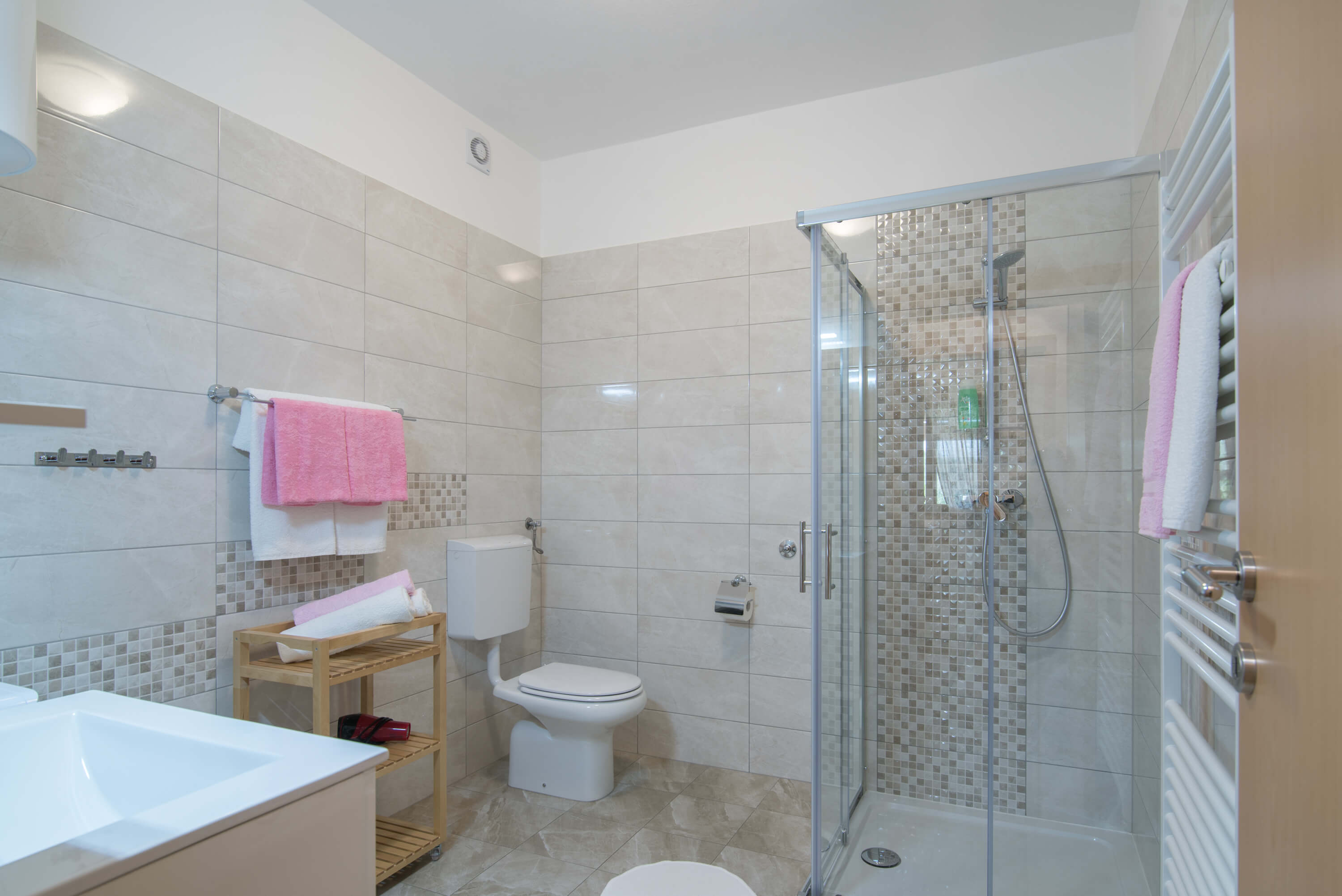 Big tiled spacious bathroom with a sink, towels, toilet, shower and radiator. Colorful towels on the hangers, radiator and towel shelf with hair dryer. Additional hangers on the wall. Toilet paper near the toilet. Shampoo in a glass shower cabin.