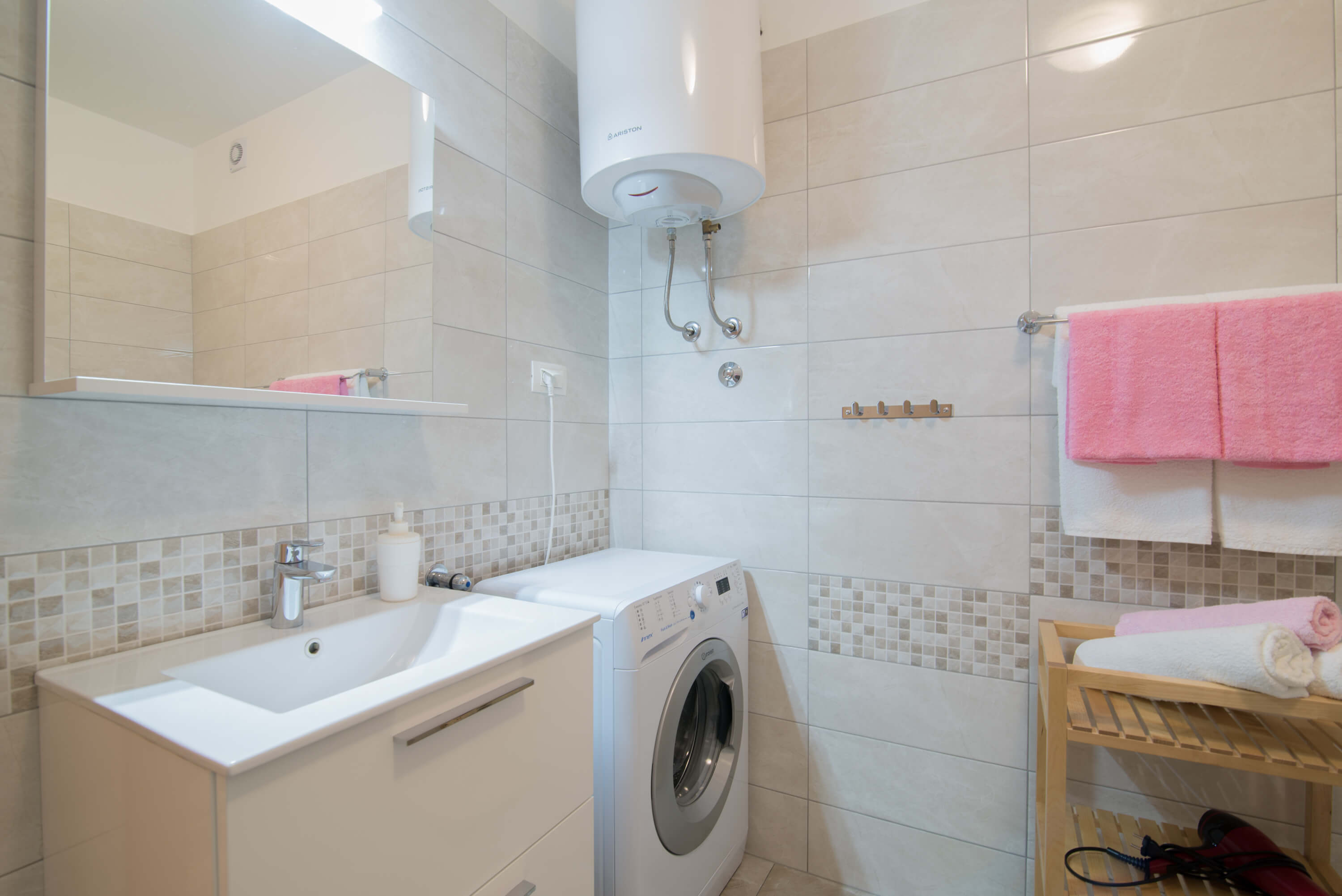 Big spacious tiled bright bathroom with appliances. Mirror above the sink with cupboards. Washing machine under the boiler. Towels in different colors on the hangers and on the towel shelf. Hair dryer on the shelf. Additional hangers on the wall.