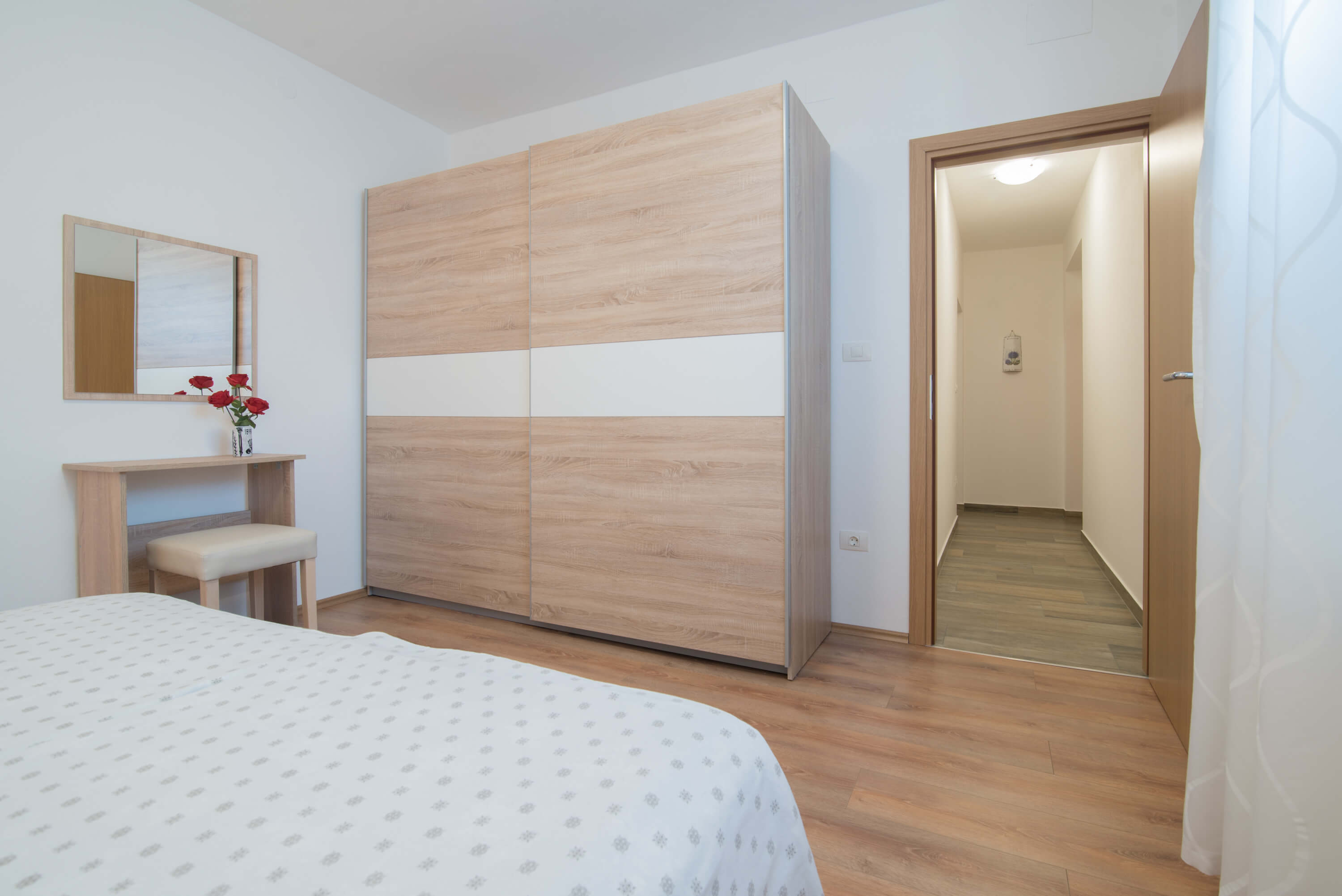 Big spacious double bed in a large bright bedroom.  Desk with a chair, flowers, and a mirror. Elegant linens on the bed. Big light brown cabinet near the door which provides a view into the hallway. Elegant curtains on the window near the door.