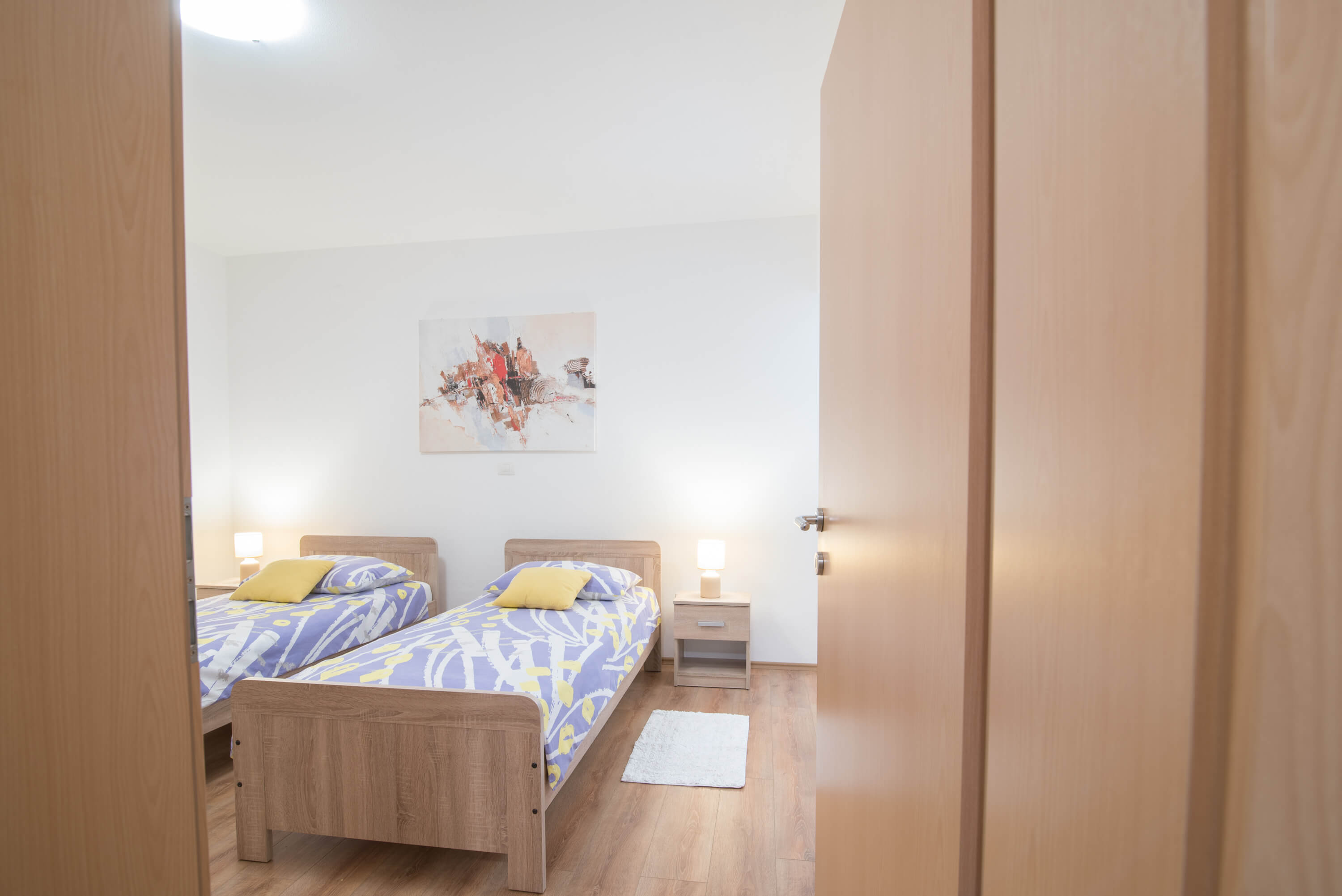 Open door entrance to a big spacious bedroom containing two single beds 90x200. Night stands and carpets next to each bed. Lamps on both night stands. Beautiful bedsheets and pillows. Painting on the wall above the beds.