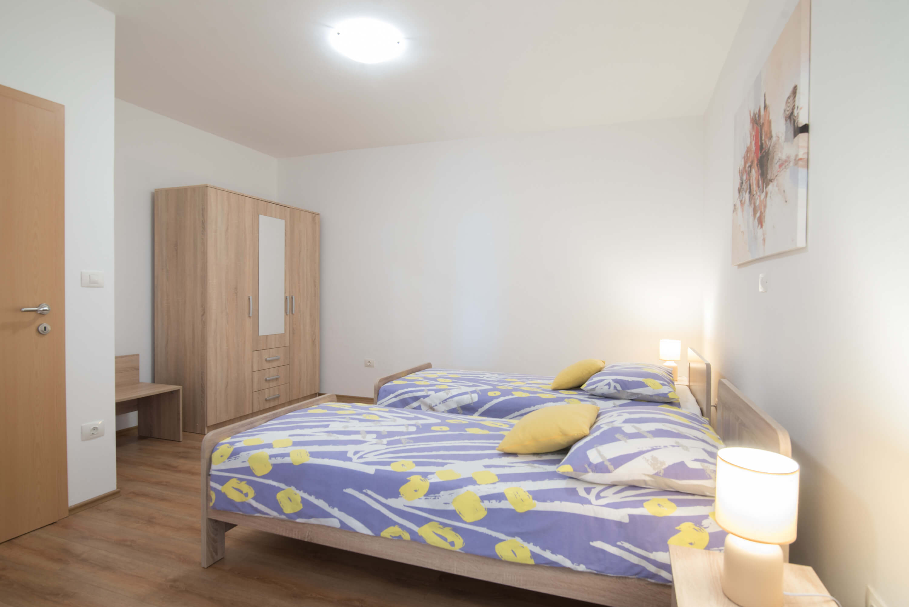 Two single beds with dimensions 90x200 in the center of a bright spacious bedroom. Colorful bedsheets and pillow linens. Lamps on night stands. Cabinet with multiple drawers and hangers alongside a luggage table. Beautiful painting on a wall.