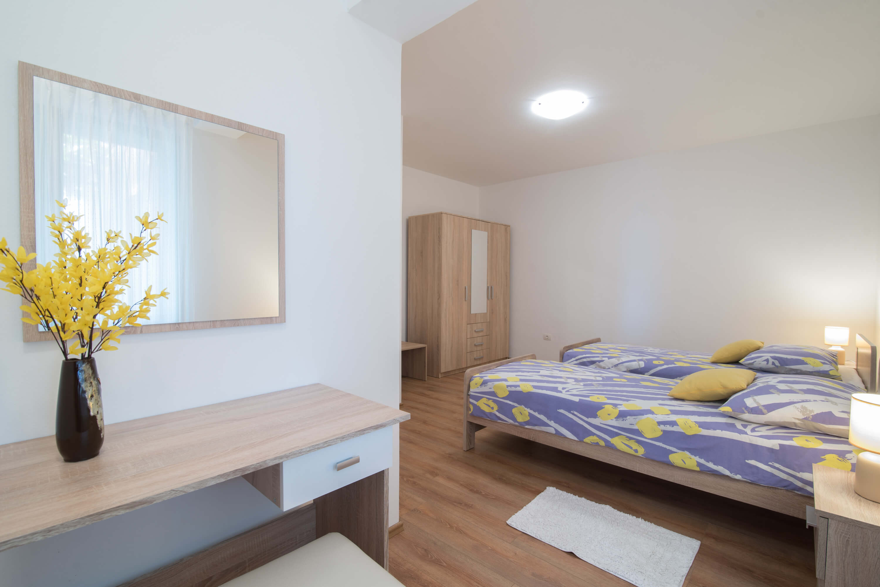 Desk with a chair, a mirror and flowers in a bedroom. Tho single beds in the center of the bedroom. Colorful linens and pillows. Carpets and night stands with lamps next to each bed. Luggage table alongside a big cabinet with drawers and hangers.
