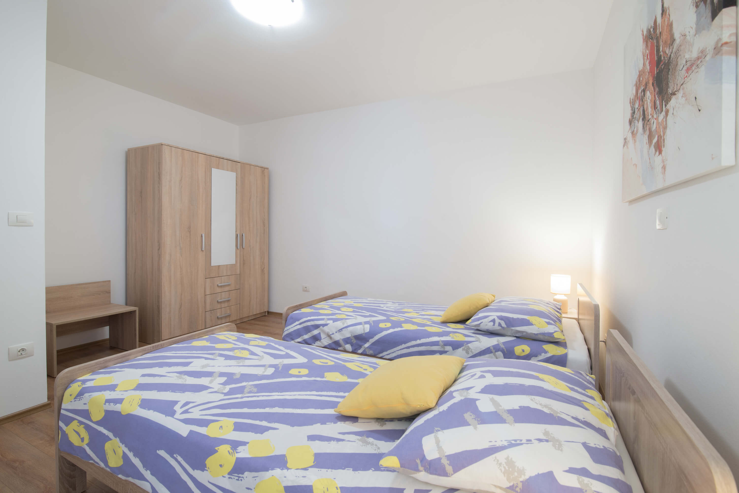 Two single beds with dimensions 90x200 in the center of a bright spacious bedroom. Colorful bedsheets and pillow linens. Lamps on night stands. Cabinet with multiple drawers and hangers alongside a luggage table. Beautiful painting on the wall.