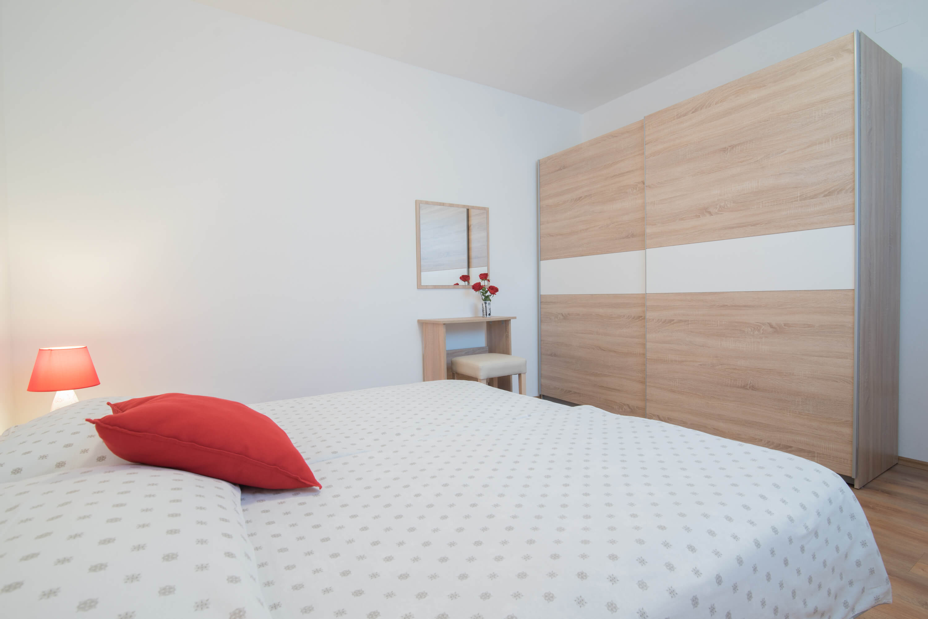 Big spacious double bed in a large bright red and white bedroom. Lamp on a night stand on the side of the bed. Desk with a chair and a mirror. Elegant linens and pillows on the bed. Painting on the wall. Big light brown cabinet.