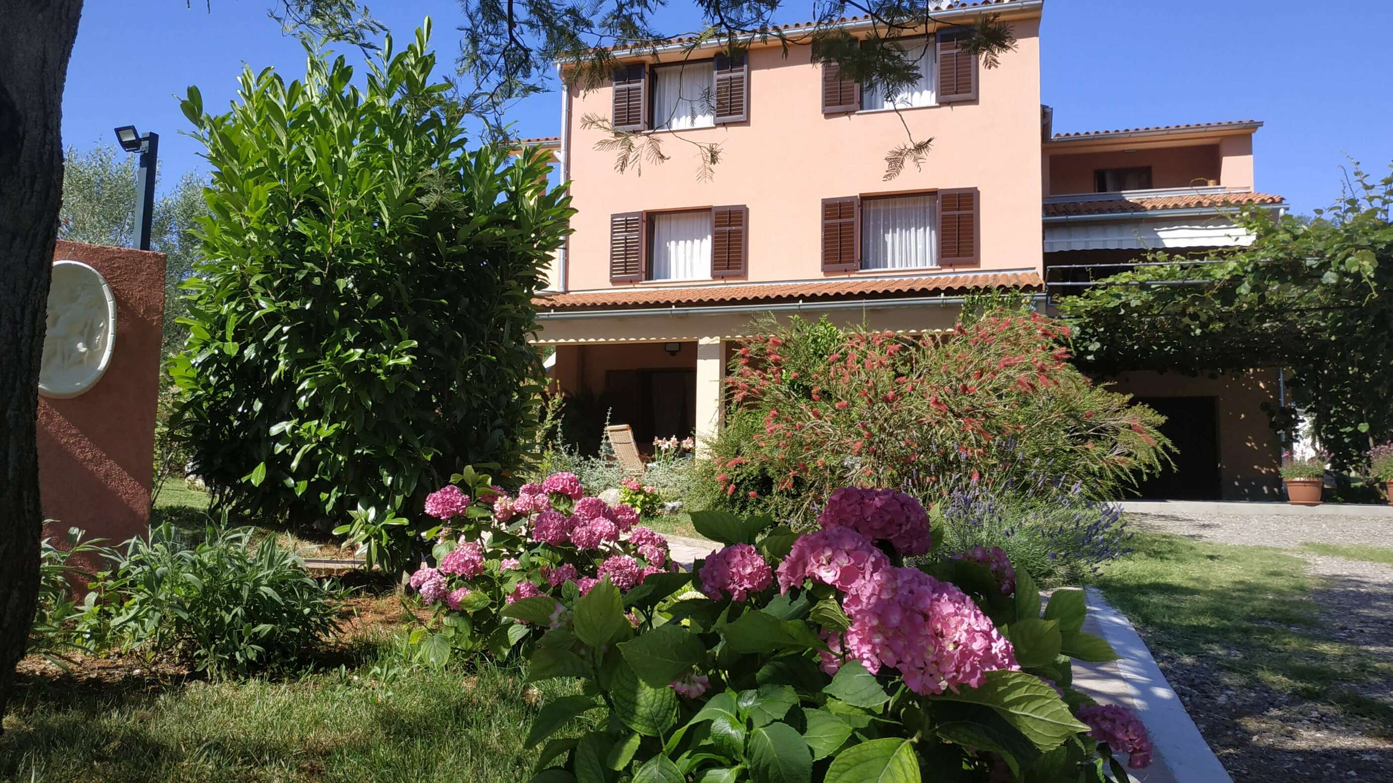 Beautiful colorful sunny garden and a lot of bautiful colorful flowers. Barbecue area with a sink. Trees and nature surrounding the garden. Pergola, house, walkway, olive growe and terrace in the background.