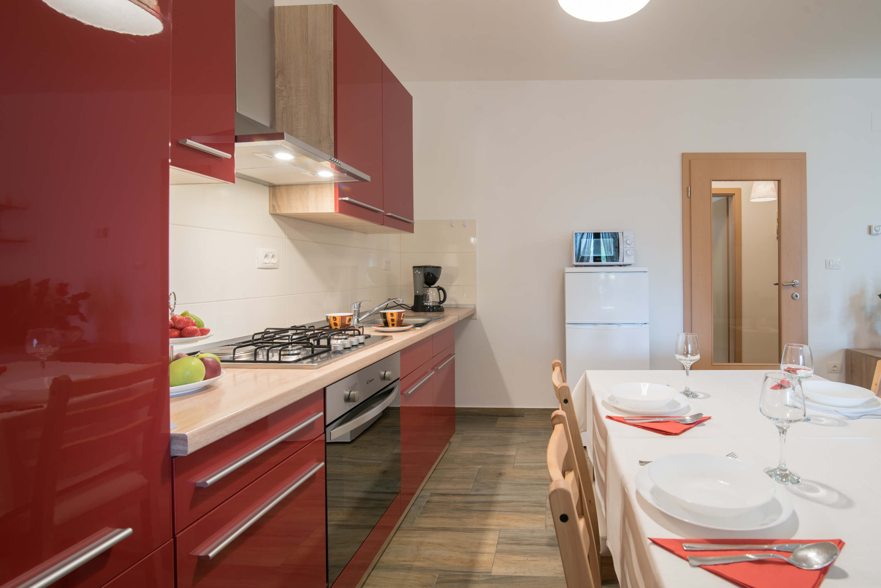 Cupboards, appliances, dining table and chairs in a big red kitchen. Set dining table surrounded by chairs. Big refrigerator and microwave. Bright kitchen workboard with cutlery and appliances including stove, oven, coffee maker and kitchen hood.