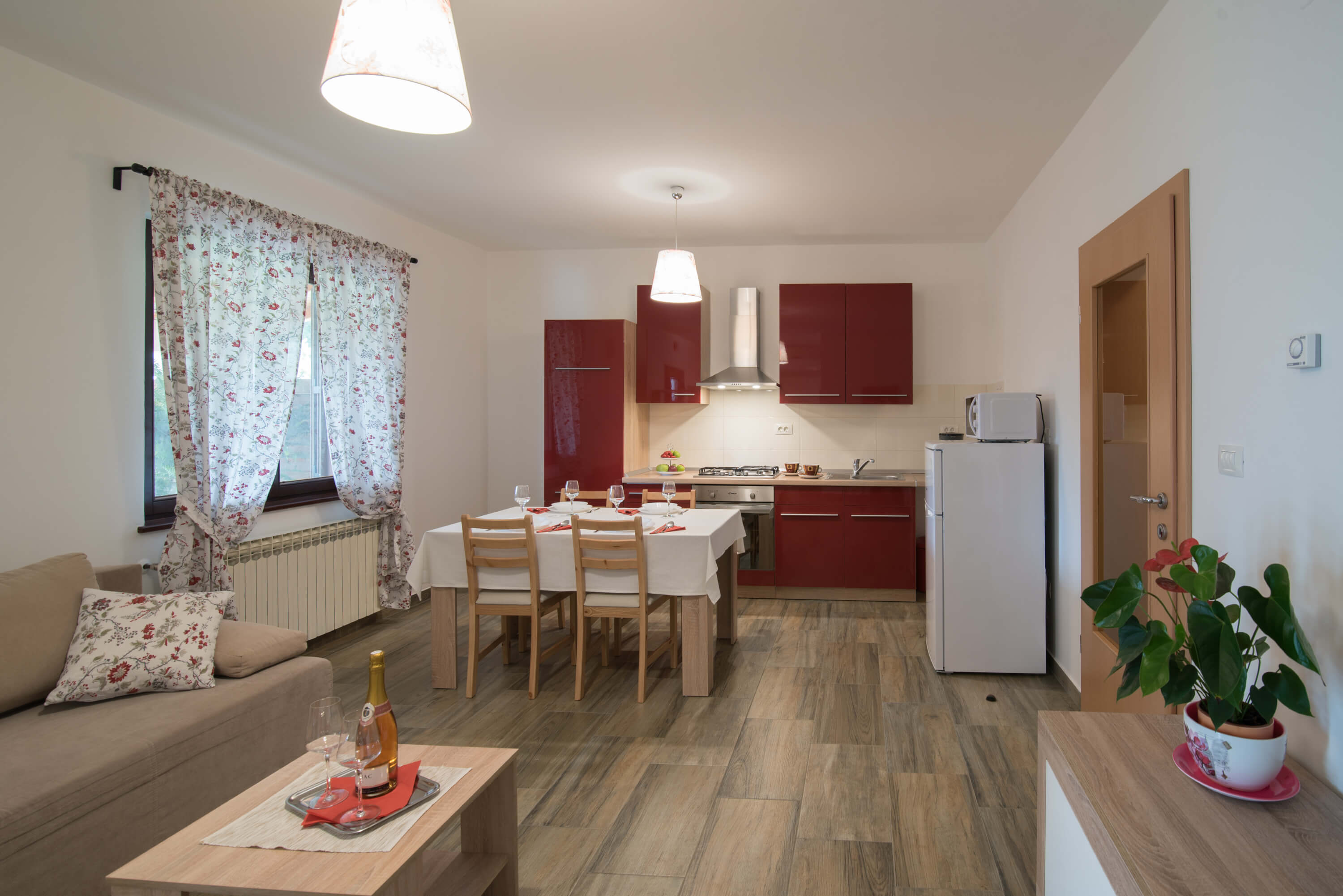 Red kitchen with all the required appliances: stove, refrigerator,  microwave and cupboards. Bright dining room with dining table and chairs and a big window next to it. Living room including coffee table and sofa.