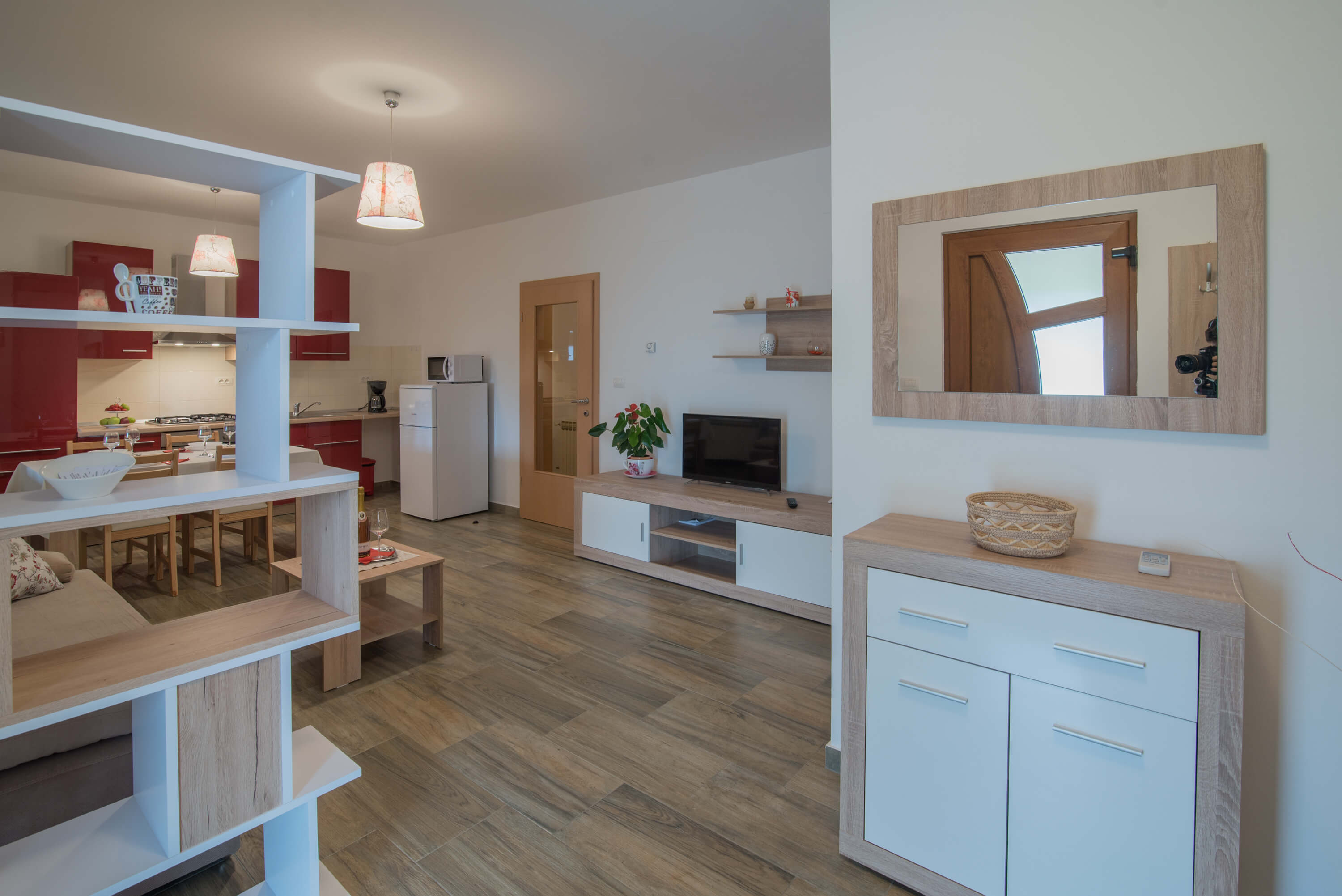 Apartment entrance with mirror, cupboard, and shelf. Living room with sofa, TV, and coffee table. Dining room with dining table and chairs. Red kitchen, including refrigerator and stove.