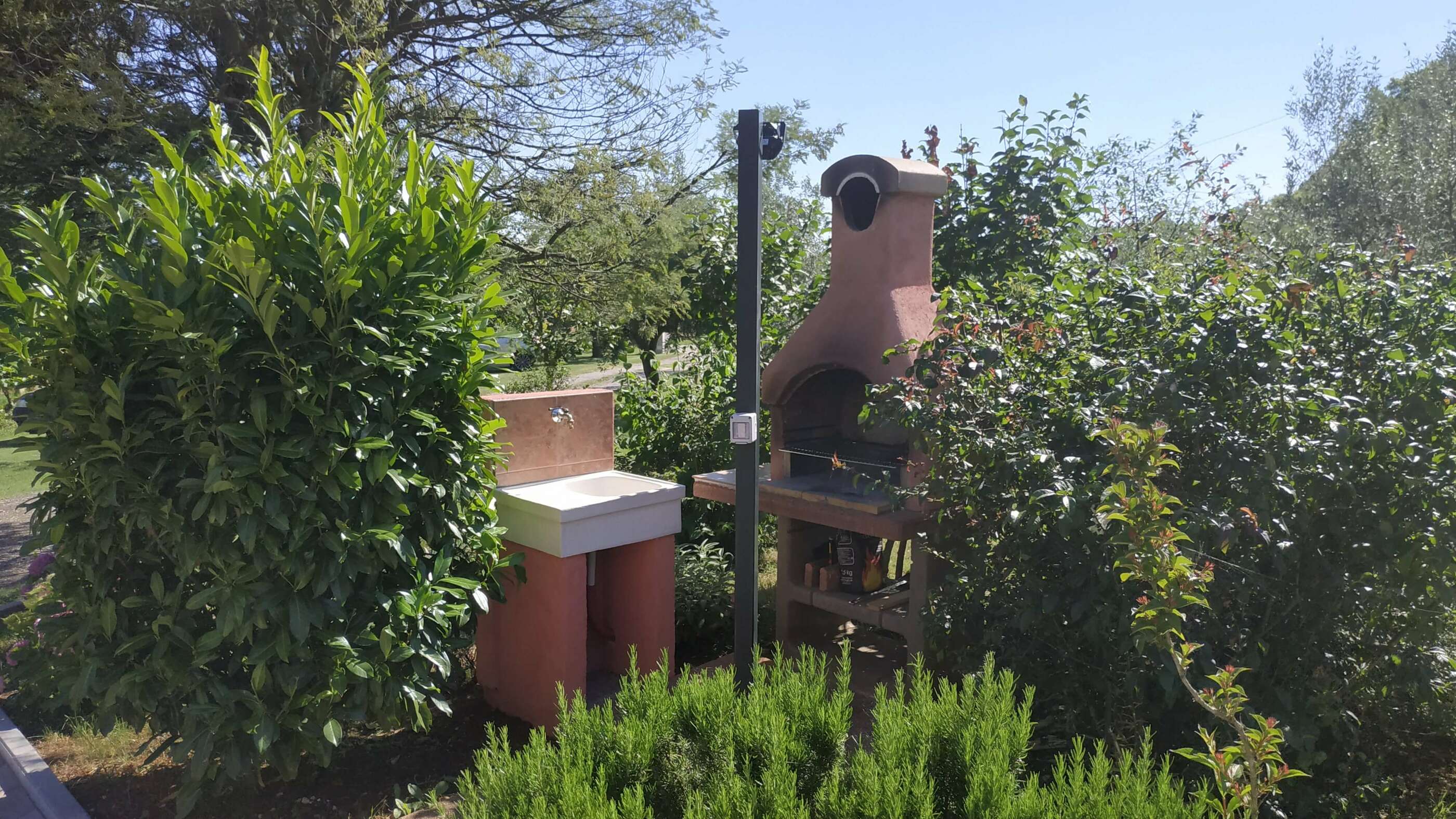 Barbecue area locted in the garden. Barbecue and sink surrounded by beatiful nature and green bushes. Tiled standing space connected to a nearby access path. Lamp light lights up the area. Sink with a faucet. Barbecue equipment under the barbecue.