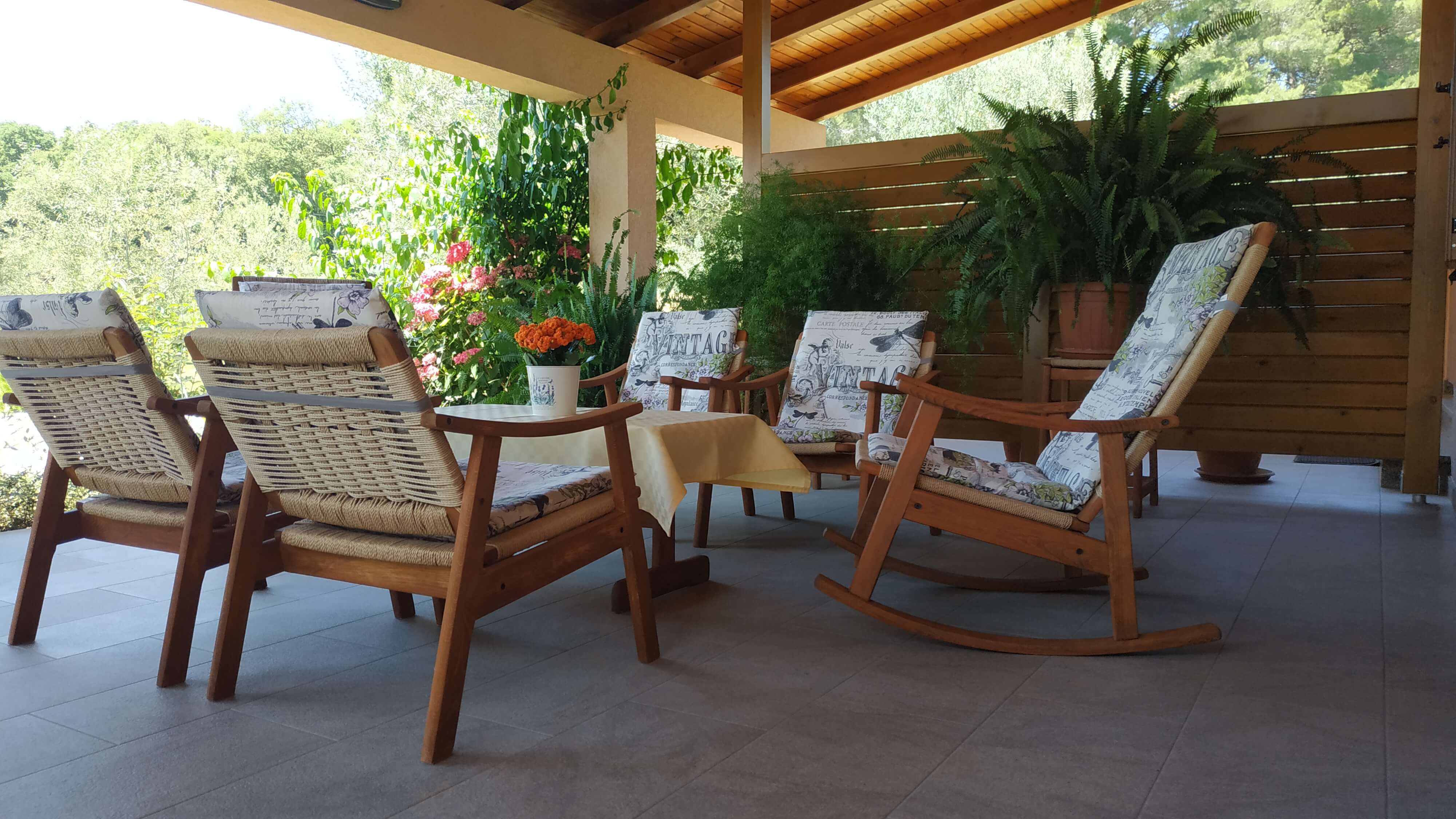 Coffee table with tablecloth and flowers on it. Armchairs next to the coffee table covered with pillows. Wooden barrier for privacy and green plants. Wooden roof and tiled floor. View into the sunny garden full of colorful flowers and plants.