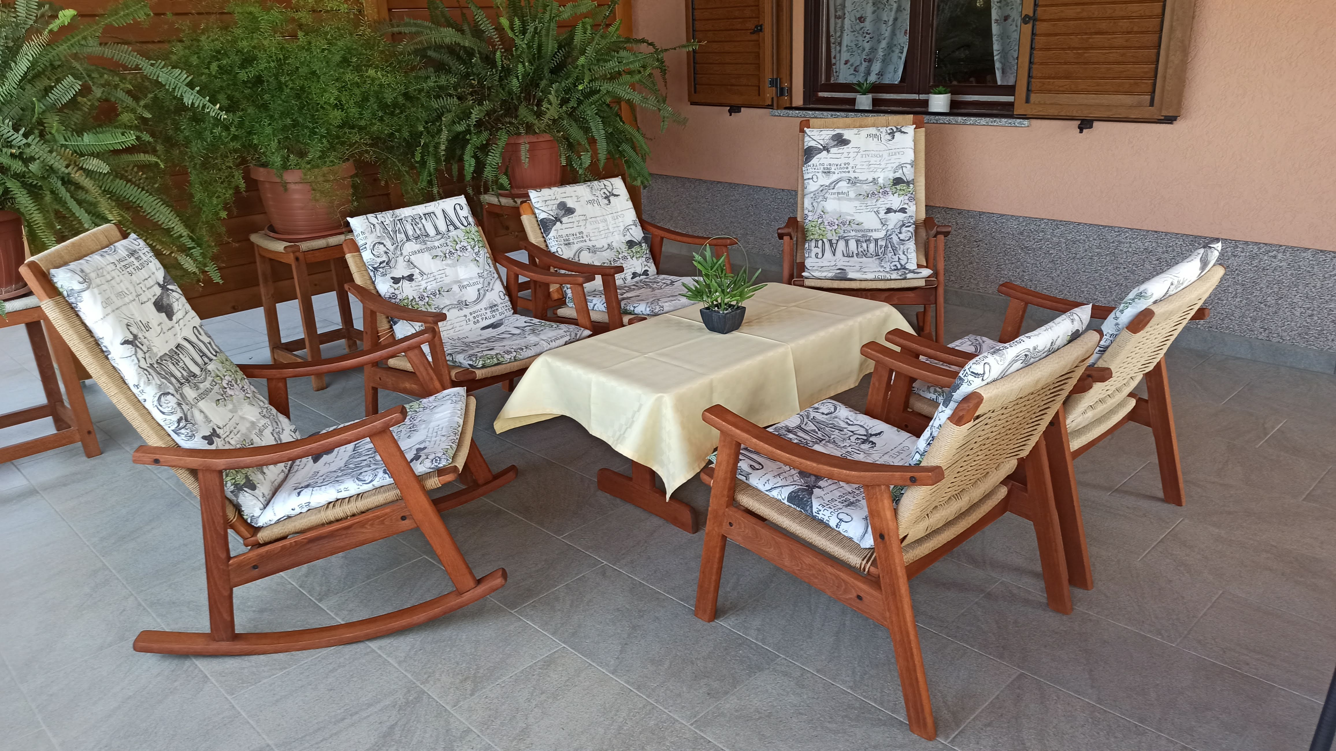 Coffee table with tablecloth and orange flowers on it. Armchairs next to the coffee table covered with pillows. Wooden barrier for privacy and green plants. Opened window on the wall with curtains and mosquito net. Elegant tiled floor.