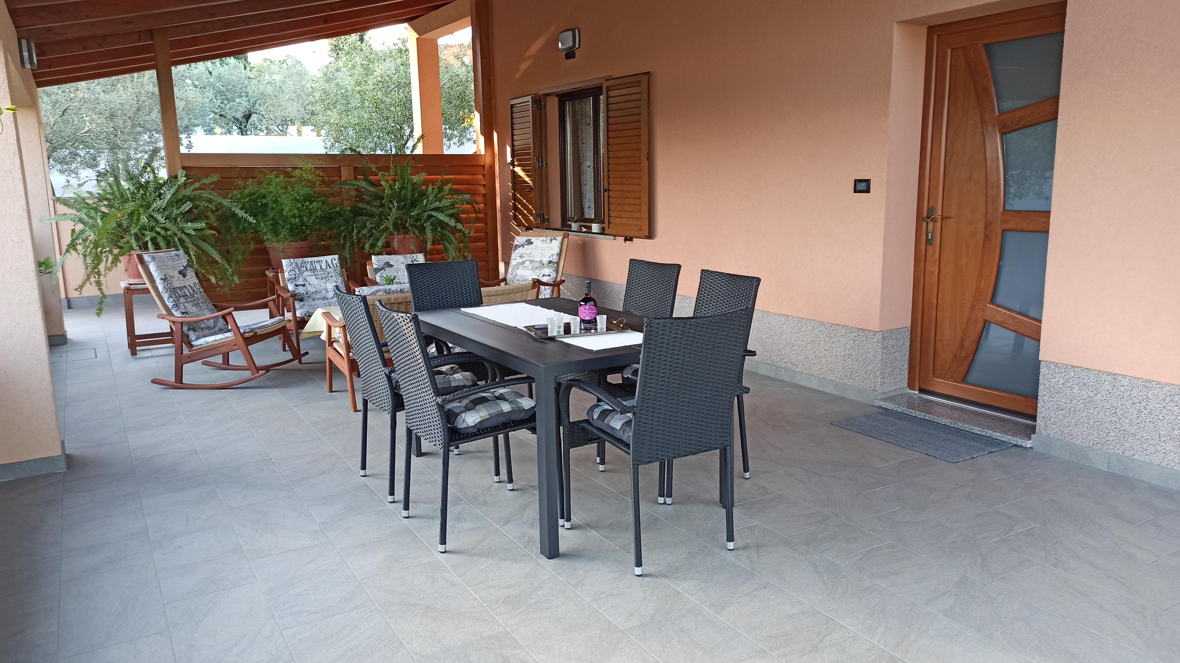 Chairs and tables on a large terrace. Dining table with plastic chairs, tablecloth and wine. Coffee table with tablecloth on it. Armchairs next to the coffee table. Entry door and a big window of the apartmene on the wall. Wooden barrier and plants.