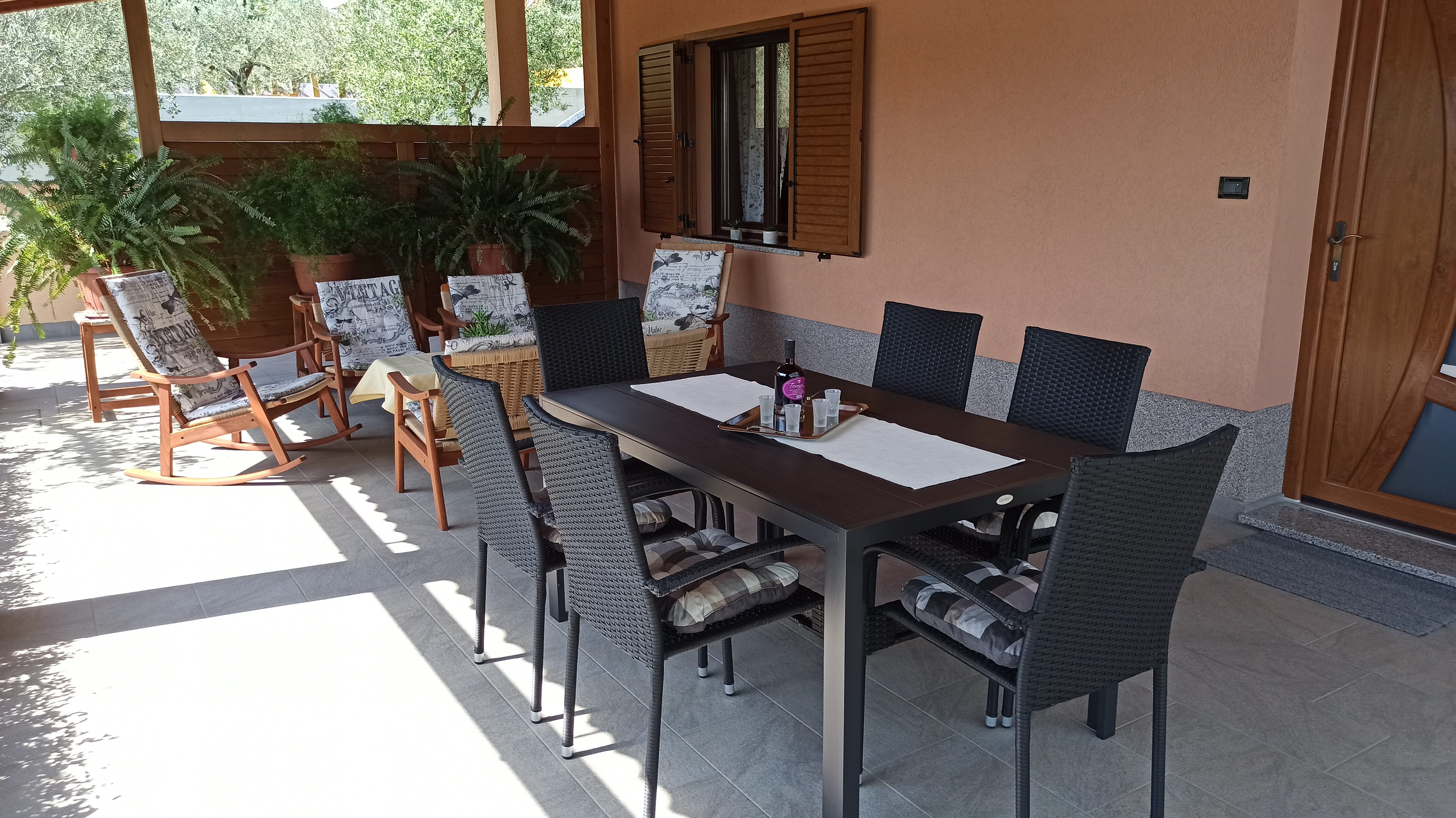 Chairs and tables on a large terrace. Dining table with plastic chairs, tablecloth and wine. Coffee table with tablecloth on it. Armchairs next to the coffee table. Entry door and a big window of the apartmene on the wall. Wooden barrier and plants.