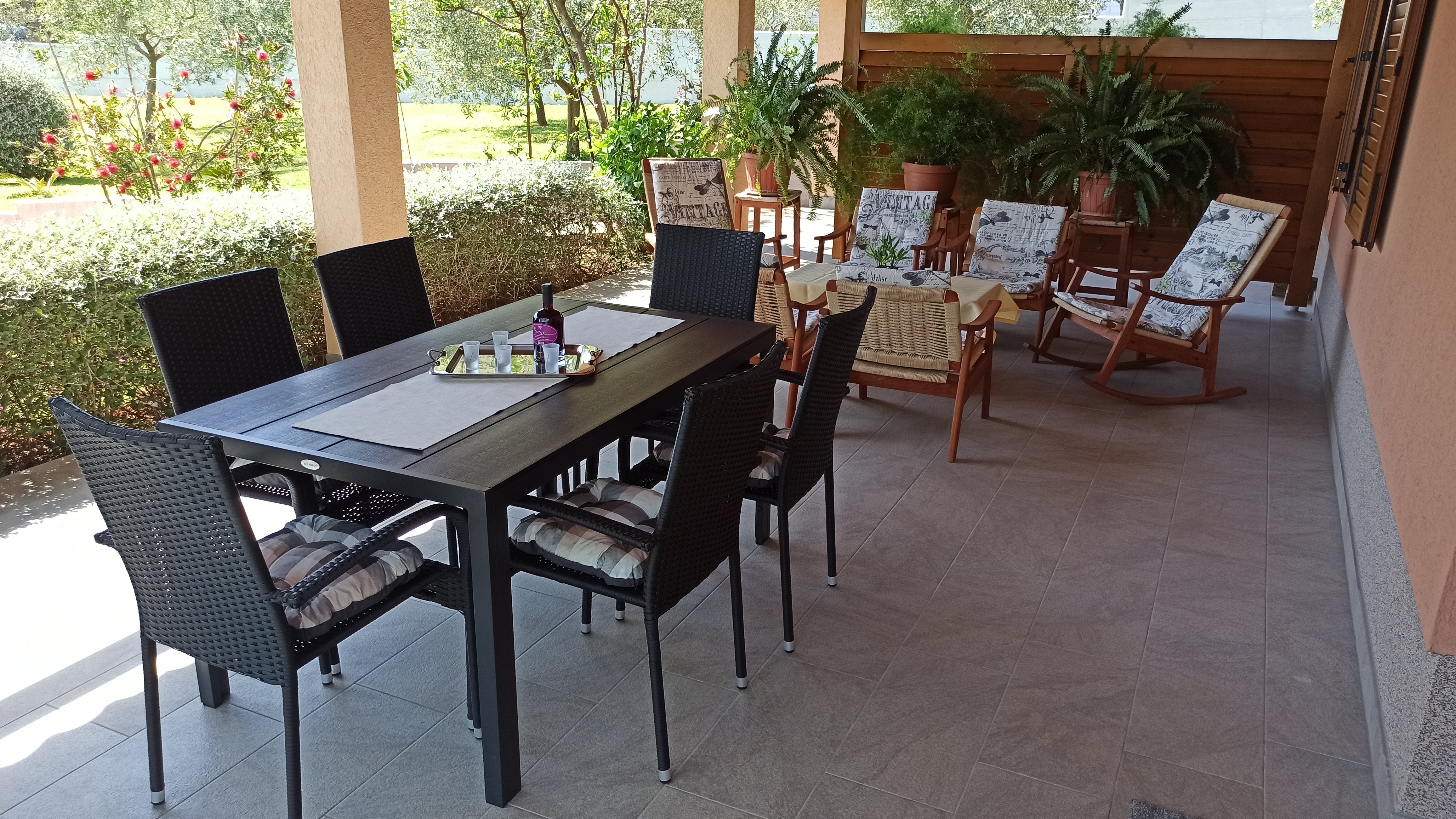 Chairs and tables on a large terrace. Dining table with plastic chairs, tablecloth and wine. Coffee table with tablecloth on it. Armchairs next to the coffee table. Big window on the wall. Wooden barrier for privacy and plants. View into the garden.