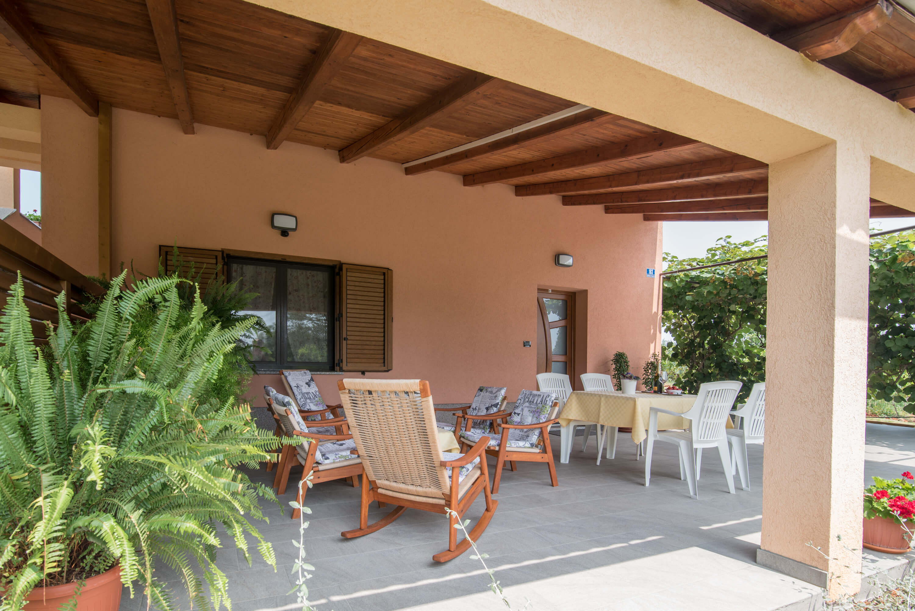 Bright, covered and tiled terrace with tables and chairs. Big dining table with chairs and coffee table surrounded by armchairs with pillows. Entrance to the apartment and a big window. Decorative plants and wooden barrier for privacy.
