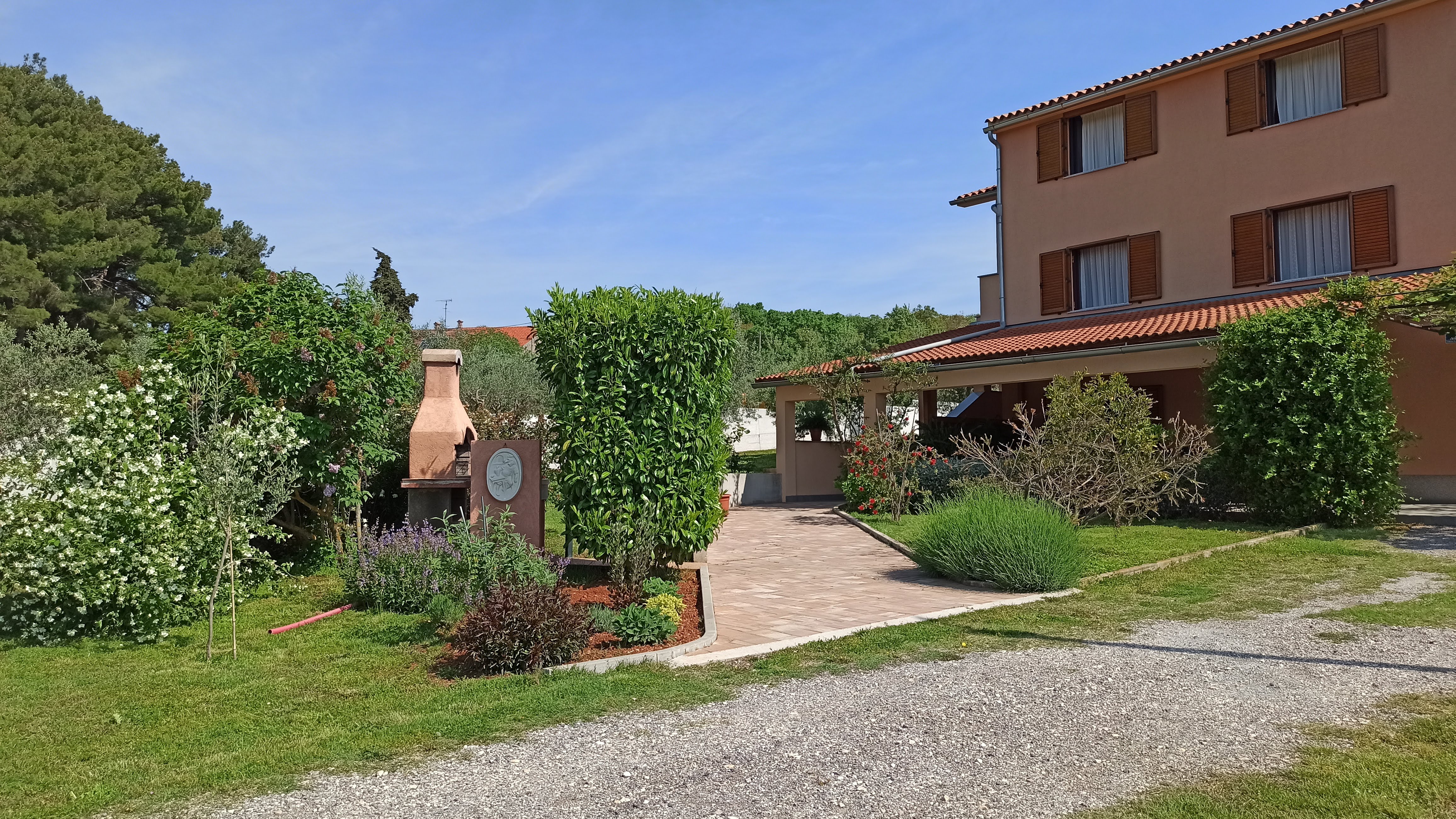 Beautiful colorful sunny garden, spacious parking space and a tiled access path. Colorful flowers filling the garden. A covered terrace and pergola in front of the apartment's entrance . Olive grove and nature in the background.