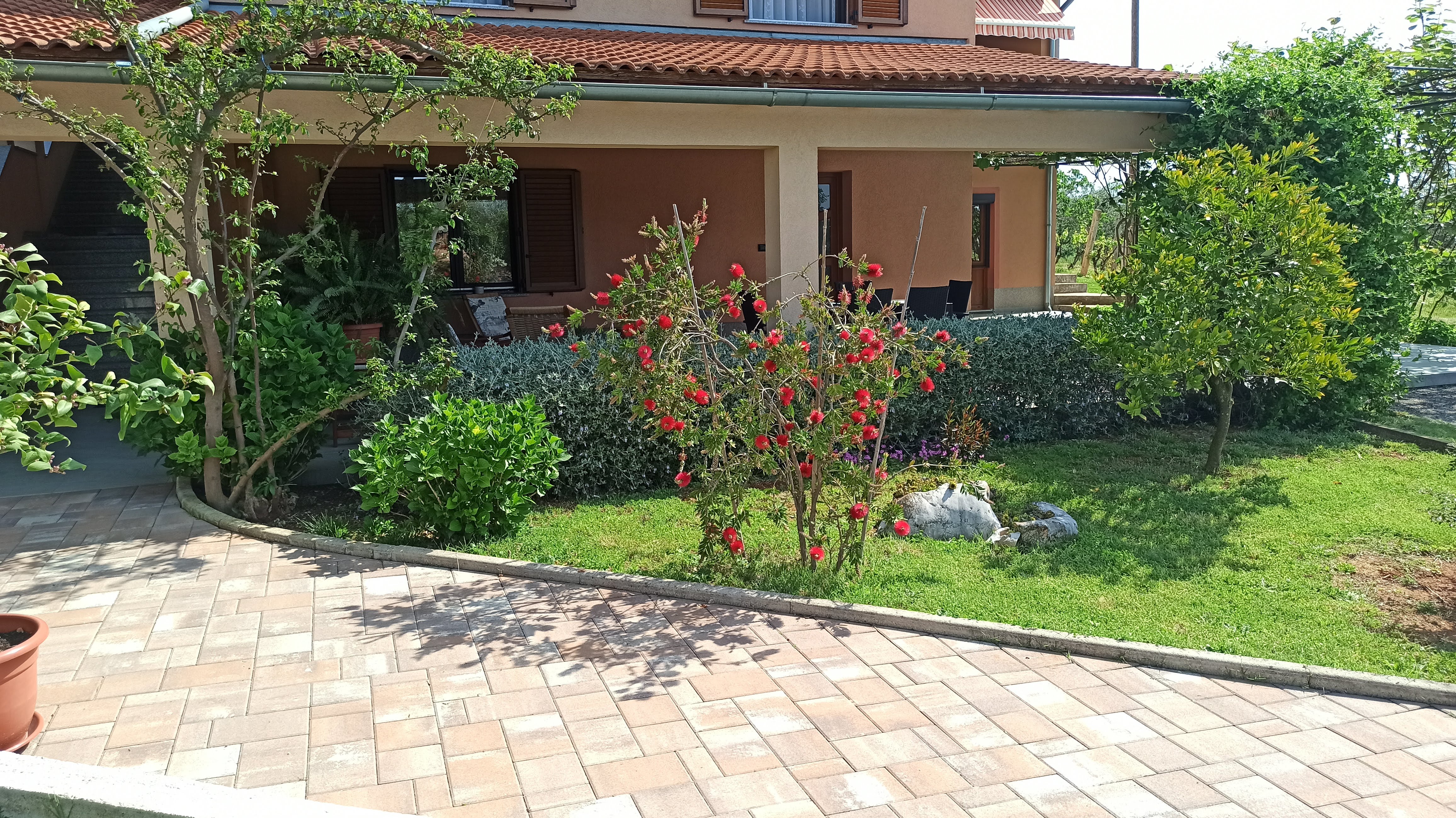 Bright, covered and tiled terrace with tables and chairs. Big dining table with chairs and coffee table surrounded by armchairs with pillows. Entrance to the apartment and a big window. Decorative plants and wooden barrier for privacy.