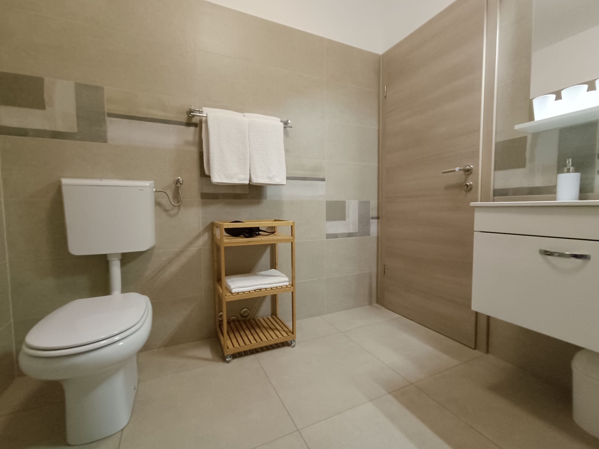 Big tiled spacious bathroom with a sink, towels, toilet, shower and radiator. Colorful towels on the hangers, radiator and towel shelf with hair dryer. Additional hangers on the wall. Toilet paper near the toilet. Shampoo in a glass shower cabin.