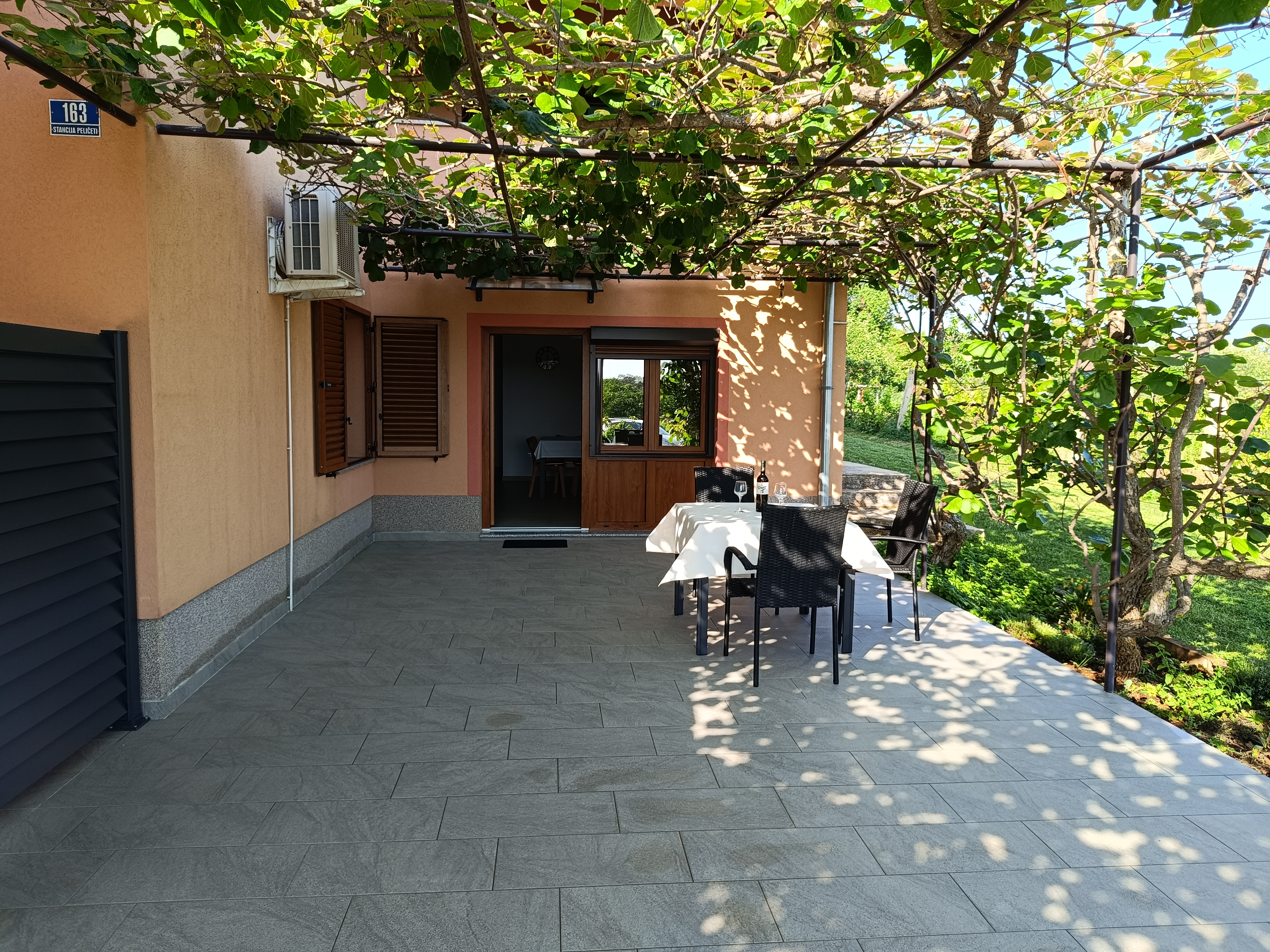 Chairs and tables on a large terrace. Dining table with chairs, tablecloth and wine. Big window on the wall. Barrier for privacy and plants. View into the garden.