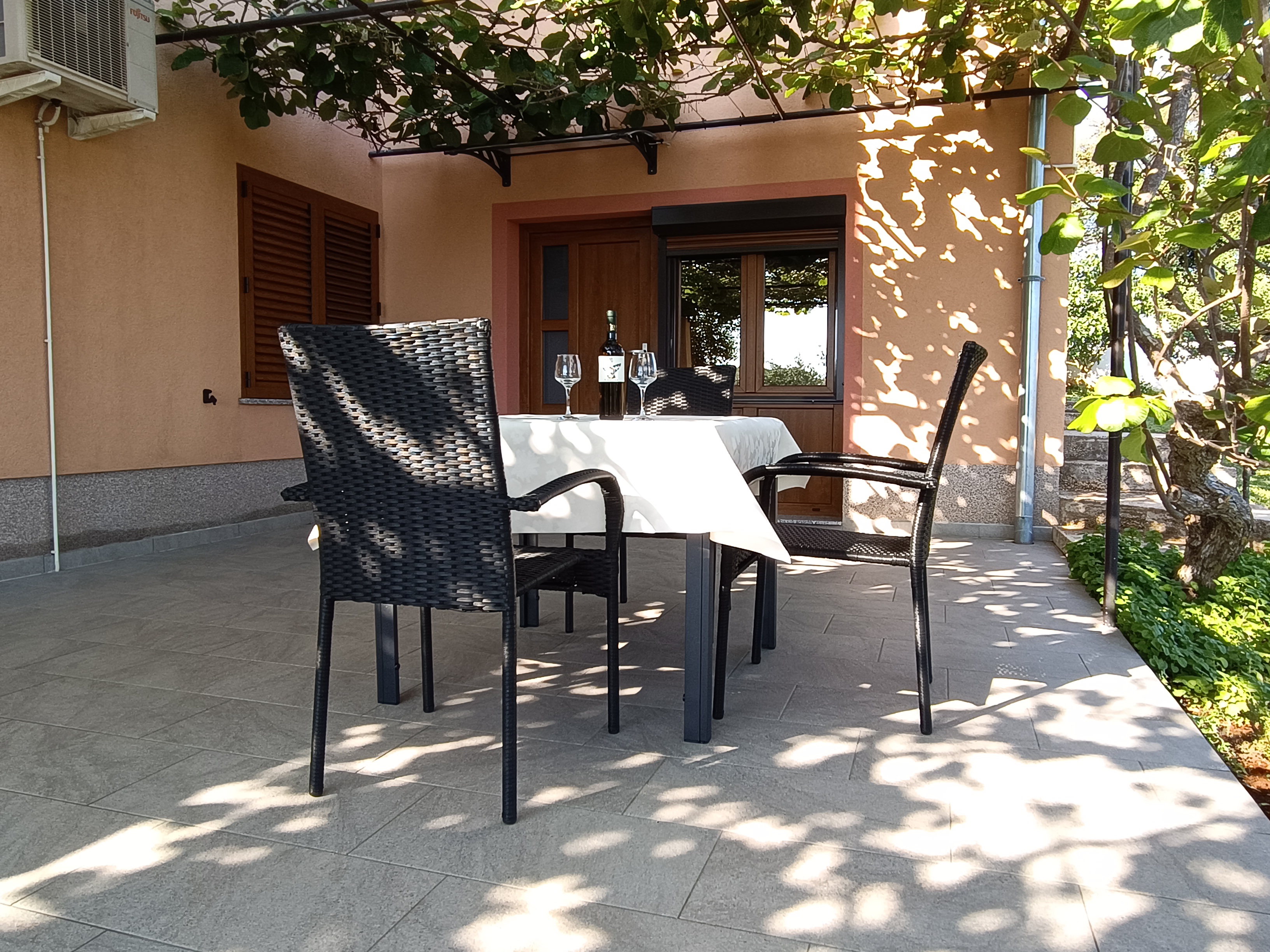 Chairs and tables on a large terrace. Dining table with chairs, tablecloth and wine. Big window on the wall. Barrier for privacy and plants. View into the garden.