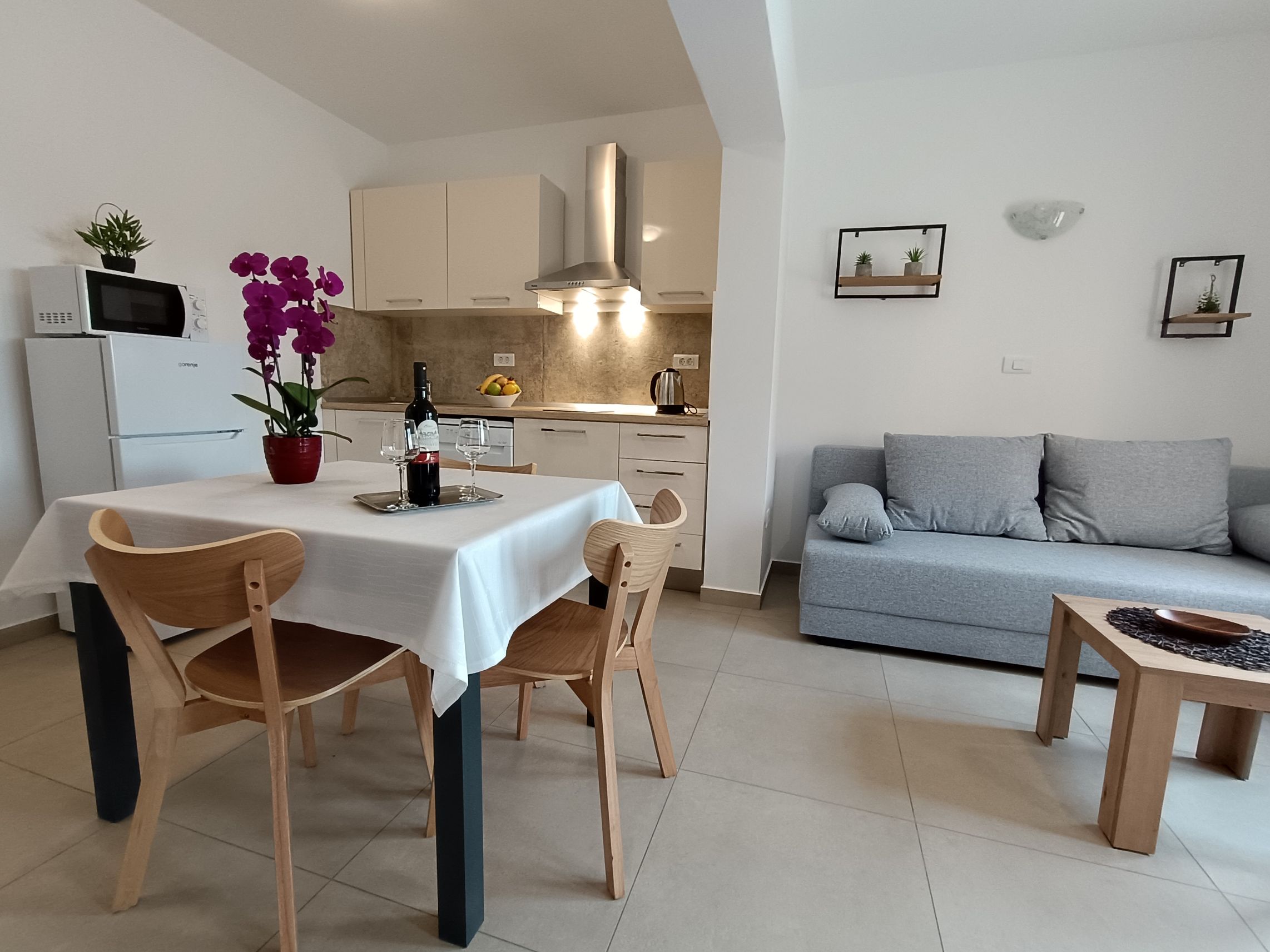 Coffee table with tablecloth and orange flowers on it. Armchairs next to the coffee table covered with pillows. Wooden barrier for privacy and green plants. Opened window on the wall with curtains and mosquito net. Elegant tiled floor.