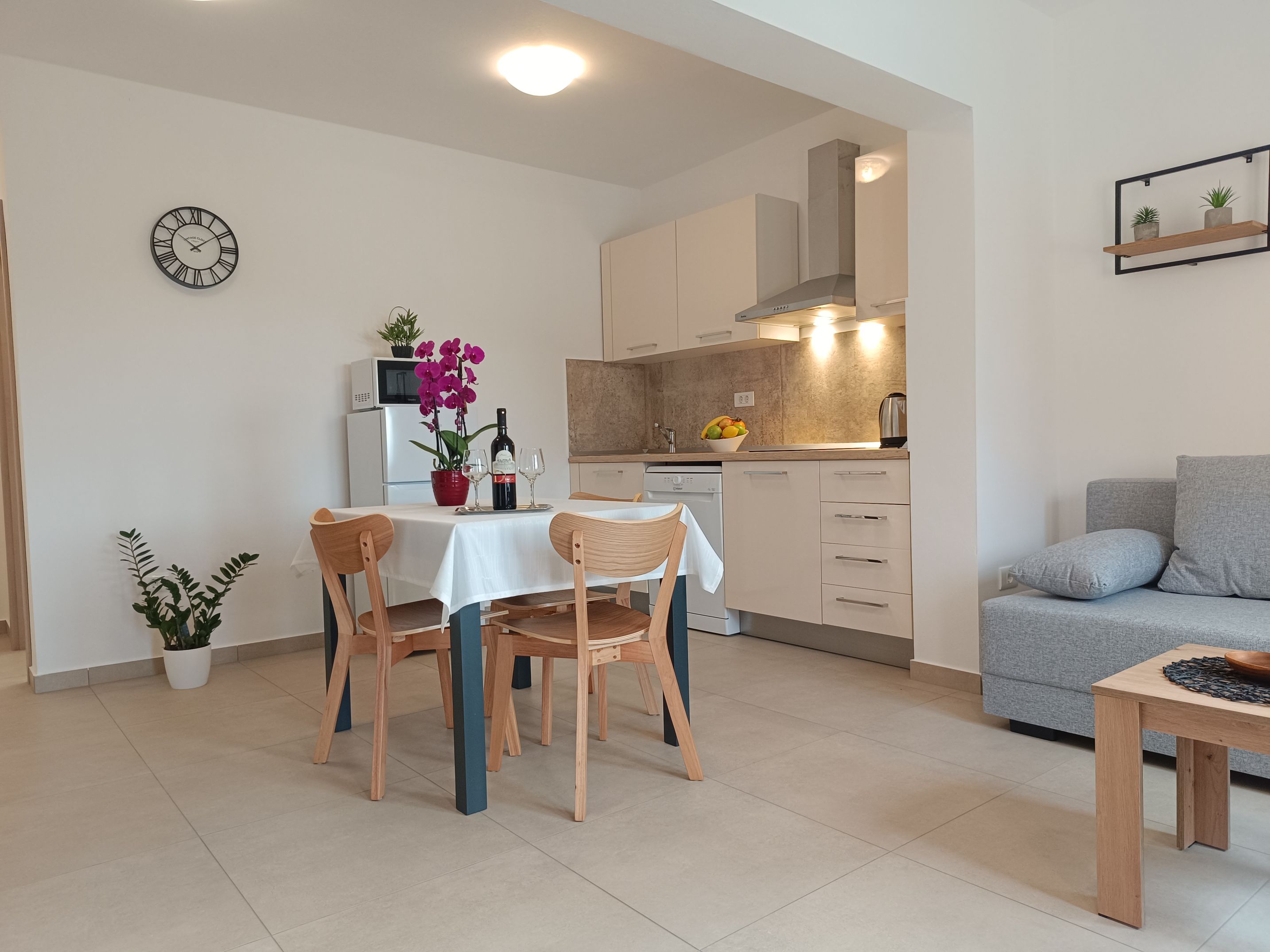 Bright, covered and tiled terrace with tables and chairs. Big dining table with chairs and coffee table surrounded by armchairs with pillows. Entrance to the apartment and a big window. Decorative plants and wooden barrier for privacy.