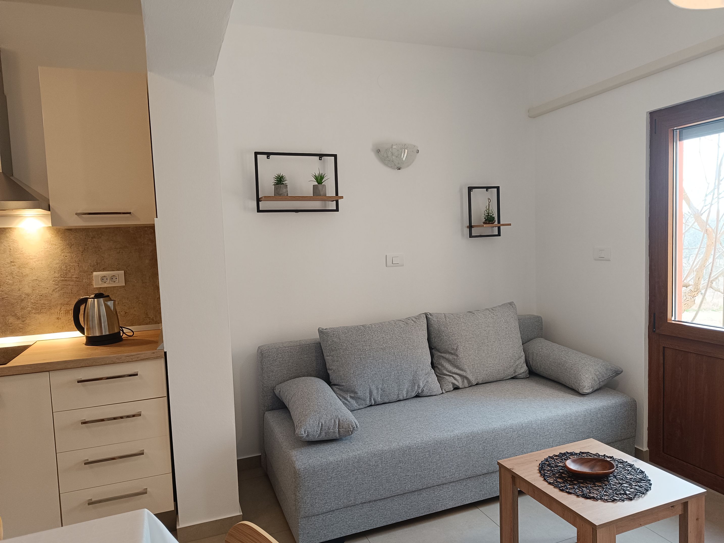 Chairs and tables on a large terrace. Dining table with plastic chairs, tablecloth and wine. Coffee table with tablecloth on it. Armchairs next to the coffee table. Entry door and a big window of the apartmene on the wall. Wooden barrier and plants.