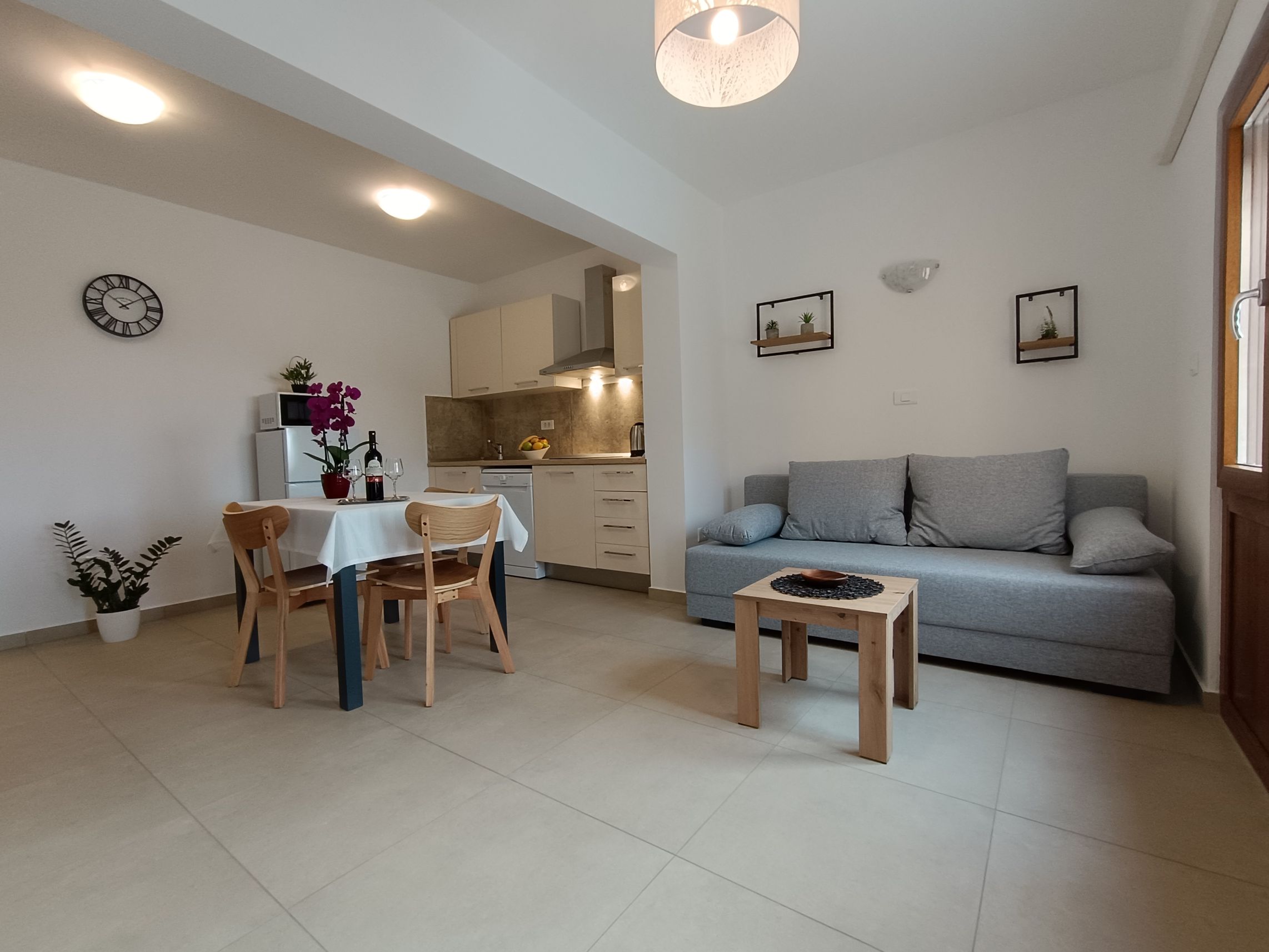 Chairs and tables on a large terrace. Dining table with plastic chairs, tablecloth and wine. Coffee table with tablecloth on it. Armchairs next to the coffee table. Big window on the wall. Wooden barrier for privacy and plants. View into the garden.